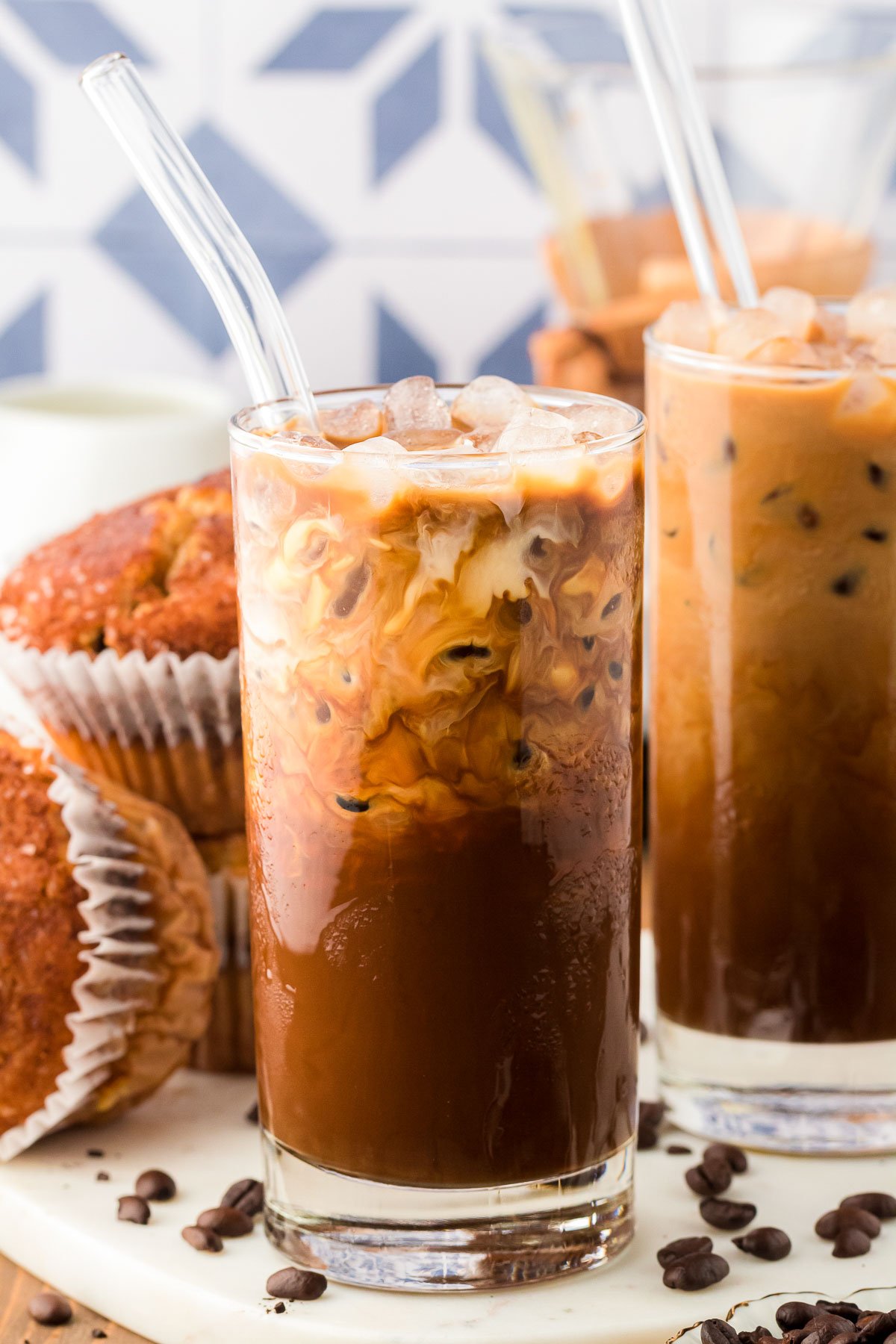 A glass of Thai Iced Coffee on a white serving board with more coffee and muffins in the background.