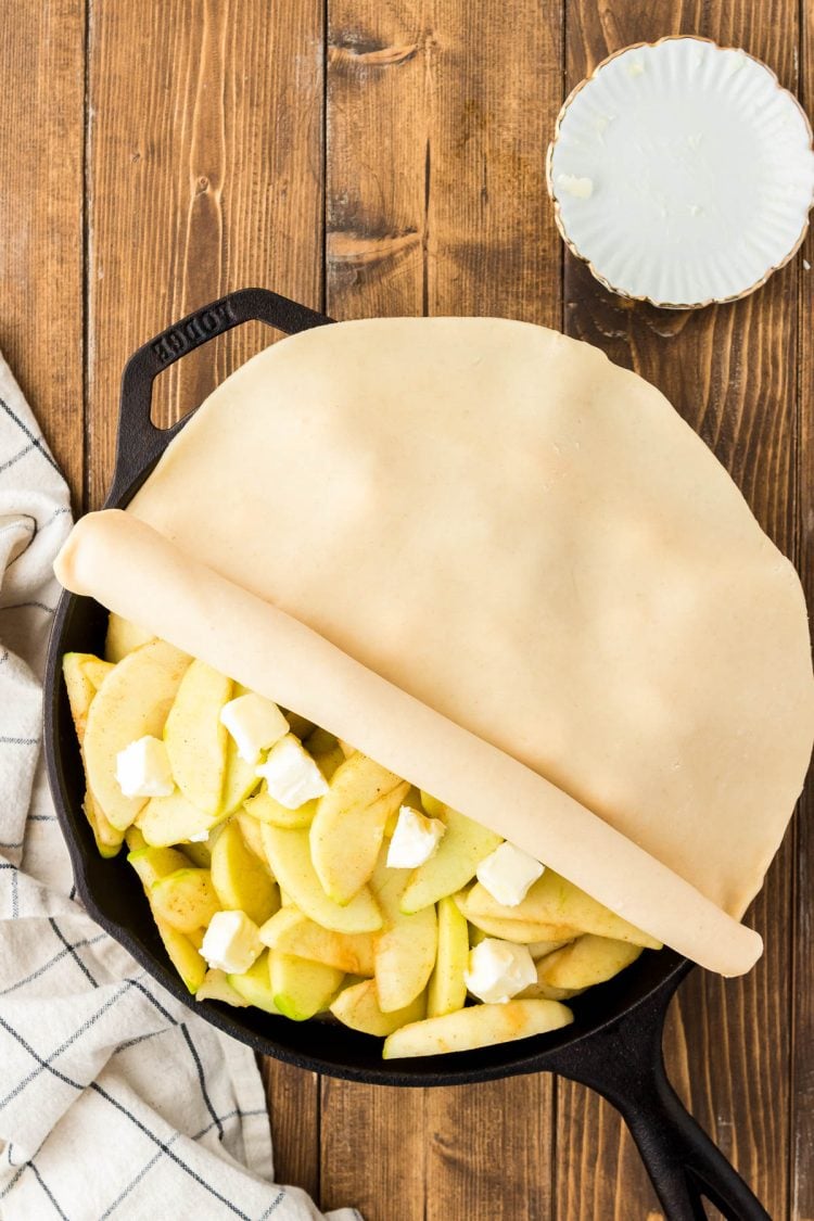 Pie crust being rolled over apples in a cast iron skillet.