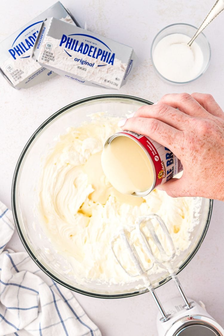 Sweetened condensed milk being poured into a mixing bowl with cream cheese and sugar.