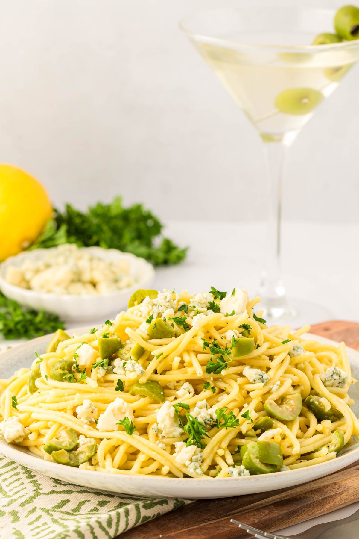 A plate of Dirty Martini Pasta on a wooden board with a martini in the background.