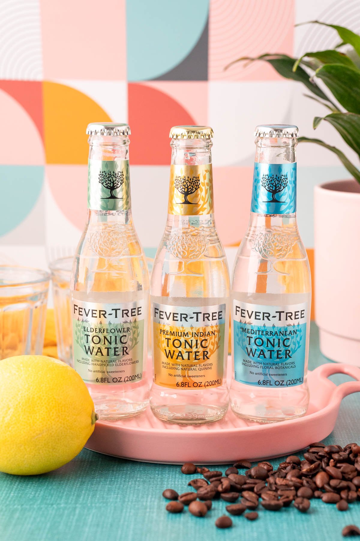 Three bottles of tonic water with a lemon and coffee beans in front of a pink serving tray. 