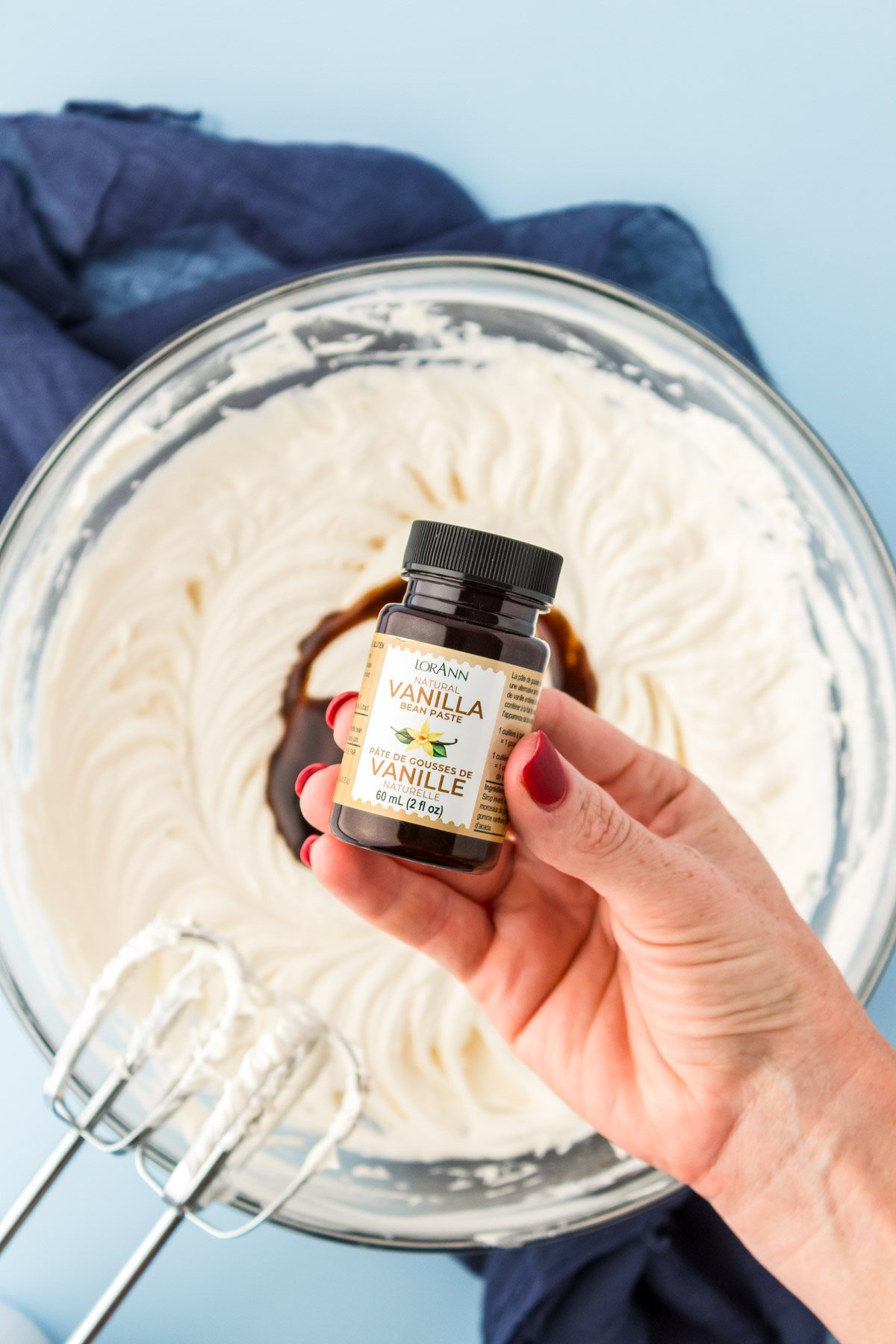 A woman's hand holding a bottle of vanilla bean paste over a bowl of vanilla frosting.