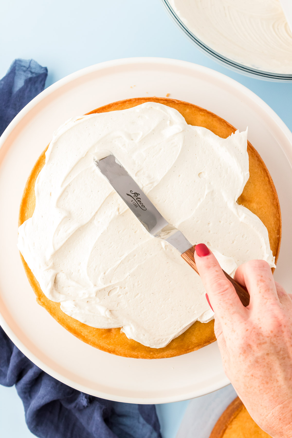 A woman's hand holding an offset spatula to spread frosting over a cake.