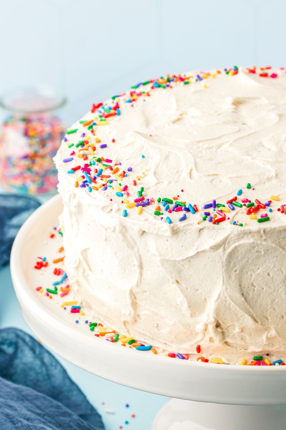 A vanilla frosted cake with rainbow sprinkles on a white cake stand.