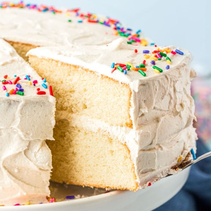 A cake server pulling a slice of vanilla cake from a white cake stand.