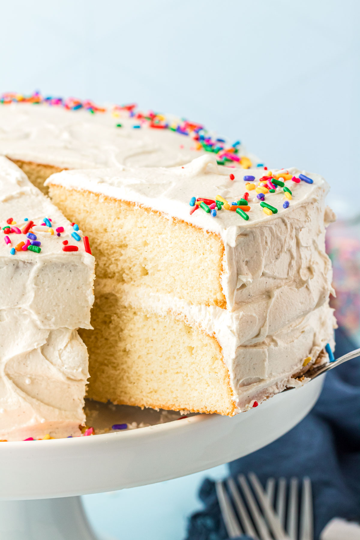 A cake server pulling a slice of vanilla cake from a white cake stand.