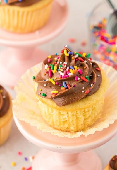 Yellow cupcake with chocolate frosting on a small cake stand with the wrapper pulled down to show texture.