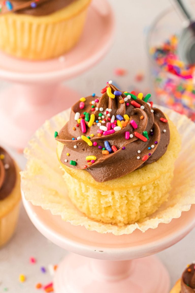 Yellow cupcake with chocolate frosting on a small cake stand with the wrapper pulled down to show texture.