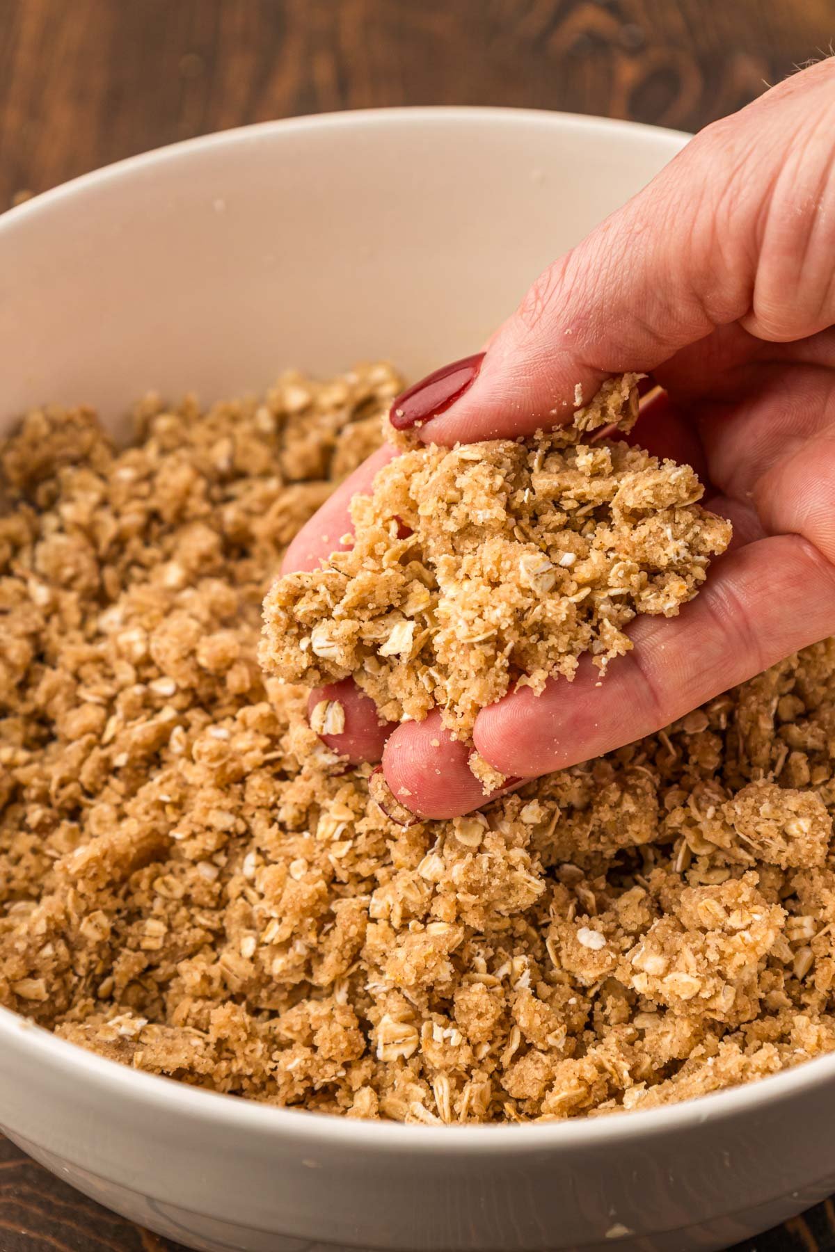 Crumble topping for apple crisp in a bowl with a woman's hand lifting some out.