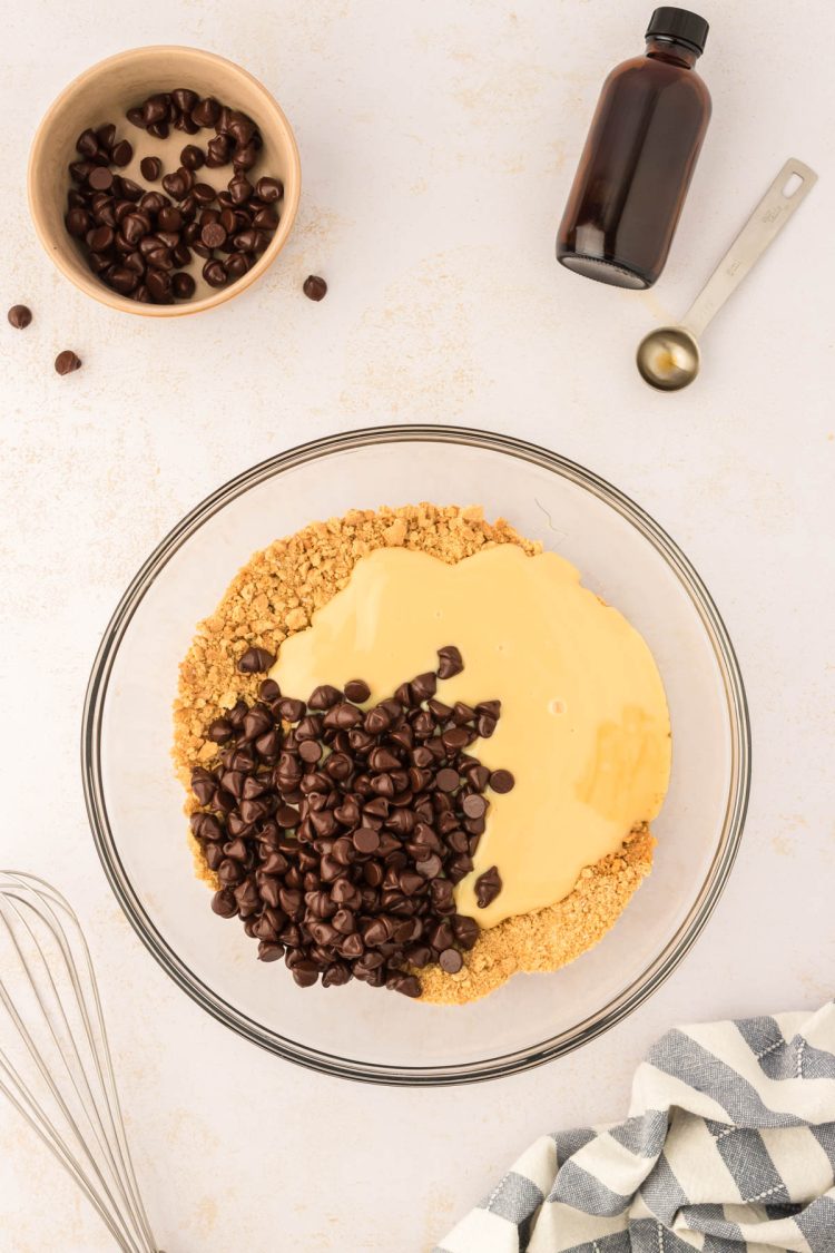 Chocolate chips and sweetened condensed milk being added to graham cracker crumbs in a bowl.