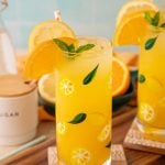 Two glasses of orangeade on coasters with citrus around them.