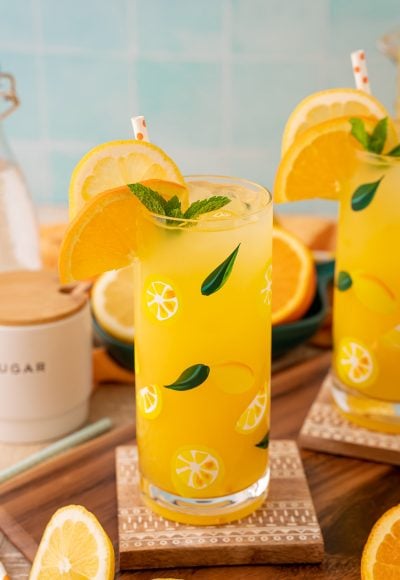 Two glasses of orangeade on coasters with citrus around them.