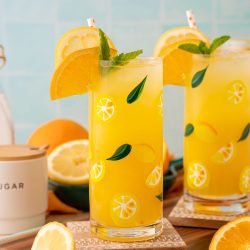 Two glasses of orangeade on coasters with citrus around them.