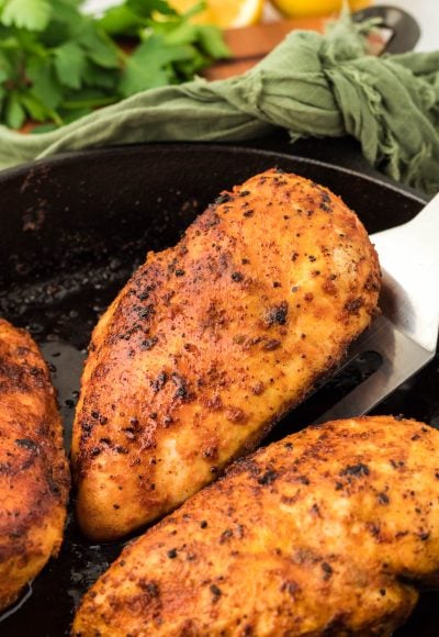 A spatula lifting a juicy chicken breast out of a cast iron skillet.