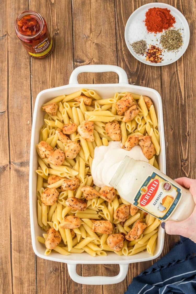 Pasta and seared chicken pieces in a casserole dish with alfredo sauce being poured on top.
