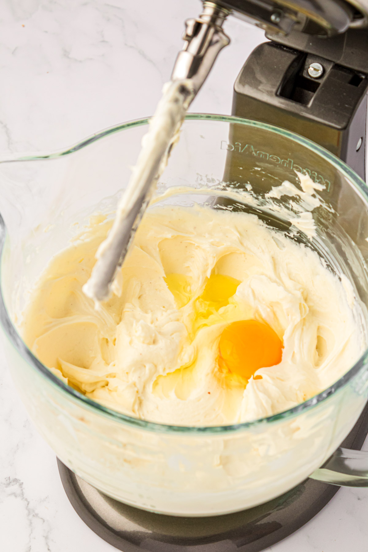 Egg being added to a stand mixer bowl with cheesecake filling.