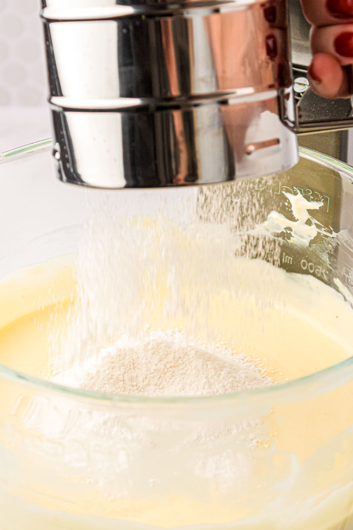 Flour being sifted into a glass mixing bowl with cheesecake filling.