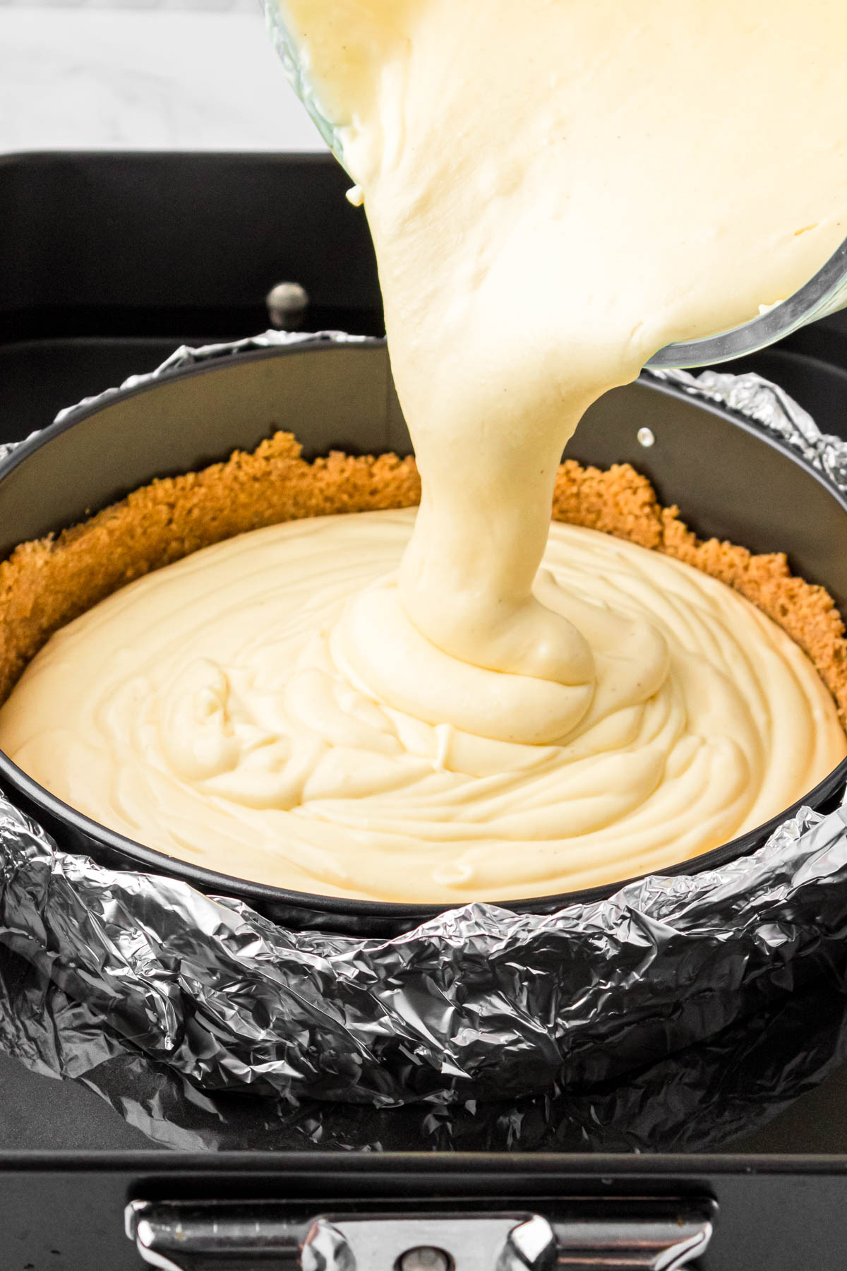 Cheesecake batter being poured into a springform pan wrapped in aluminum foil in a water bath.