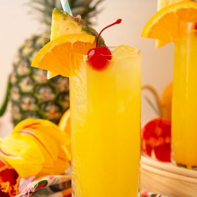 A tall glass of pineapple punch garnished with a cherry, orange slice, and pineapple wedge.