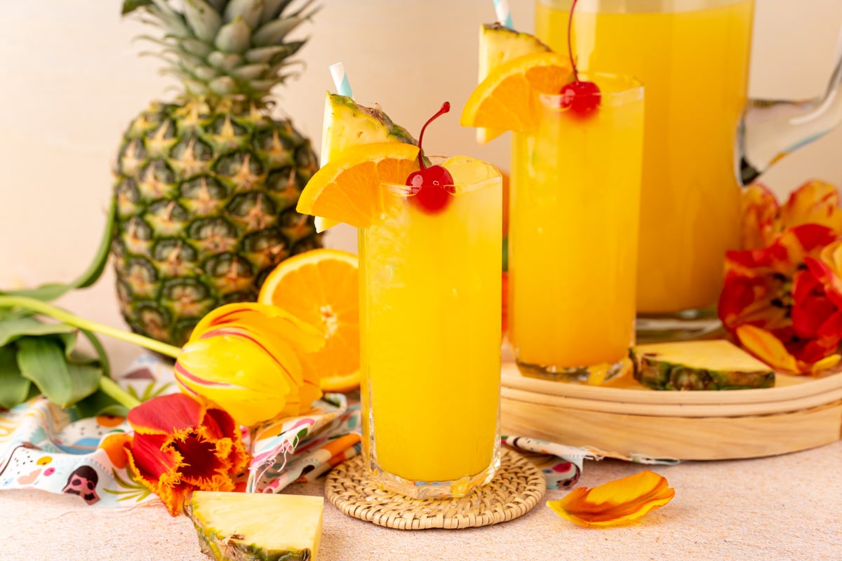 Two glasses and a pitcher of pineapple punch on a beach backdrop.