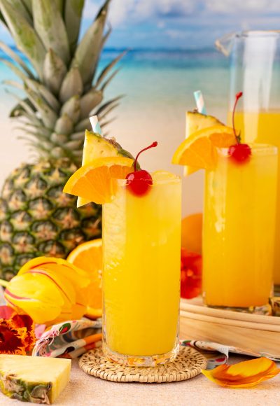 Two glasses and a pitcher of pineapple punch on a beach backdrop.