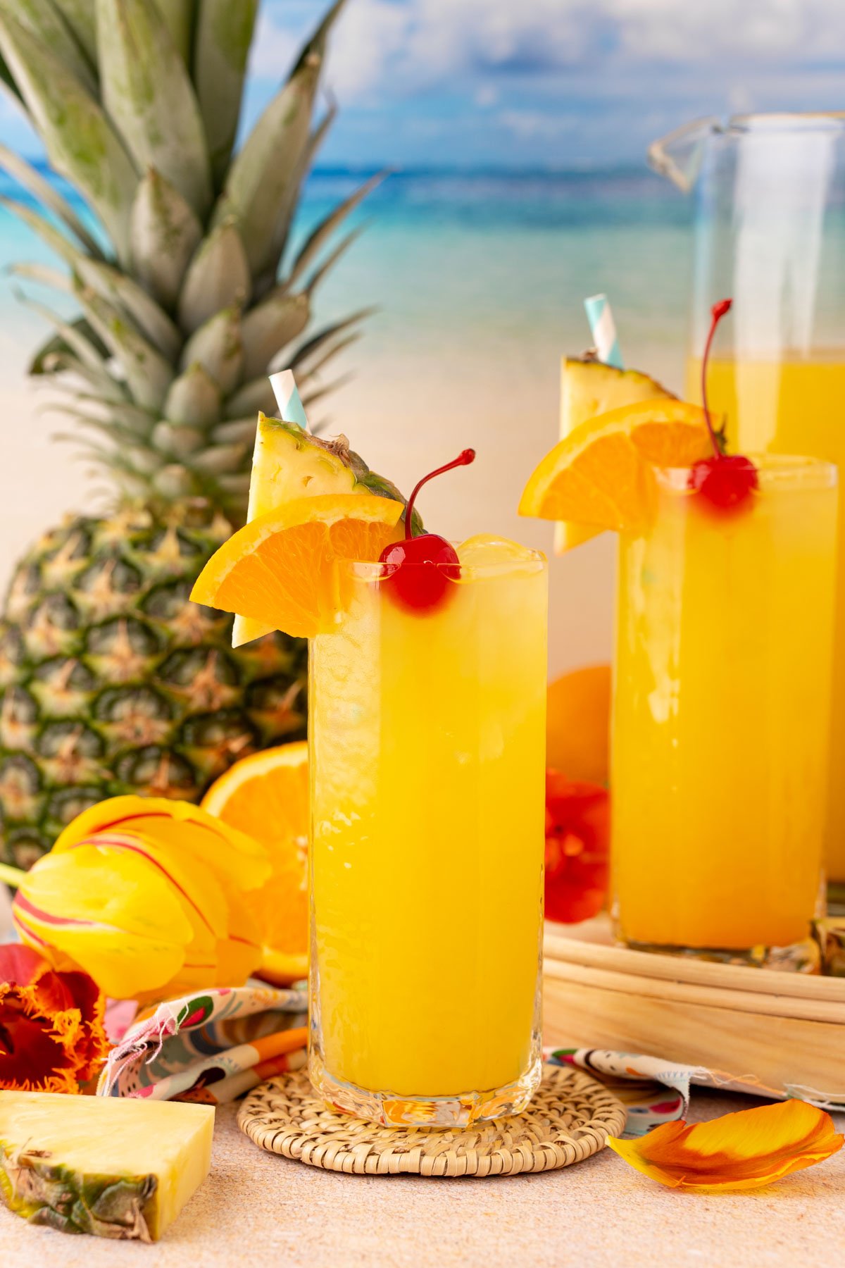 Two glasses and a pitcher of pineapple punch on a beach backdrop.