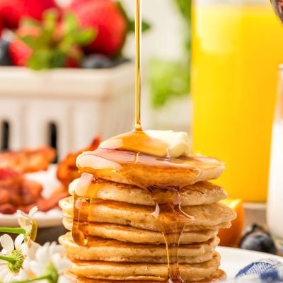 A stack of silver dollar pancakes on a white plate with syrup being poured over the top of them.