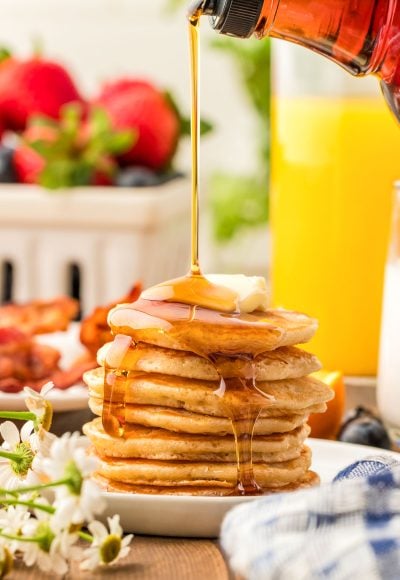 A stack of silver dollar pancakes on a white plate with syrup being poured over the top of them.