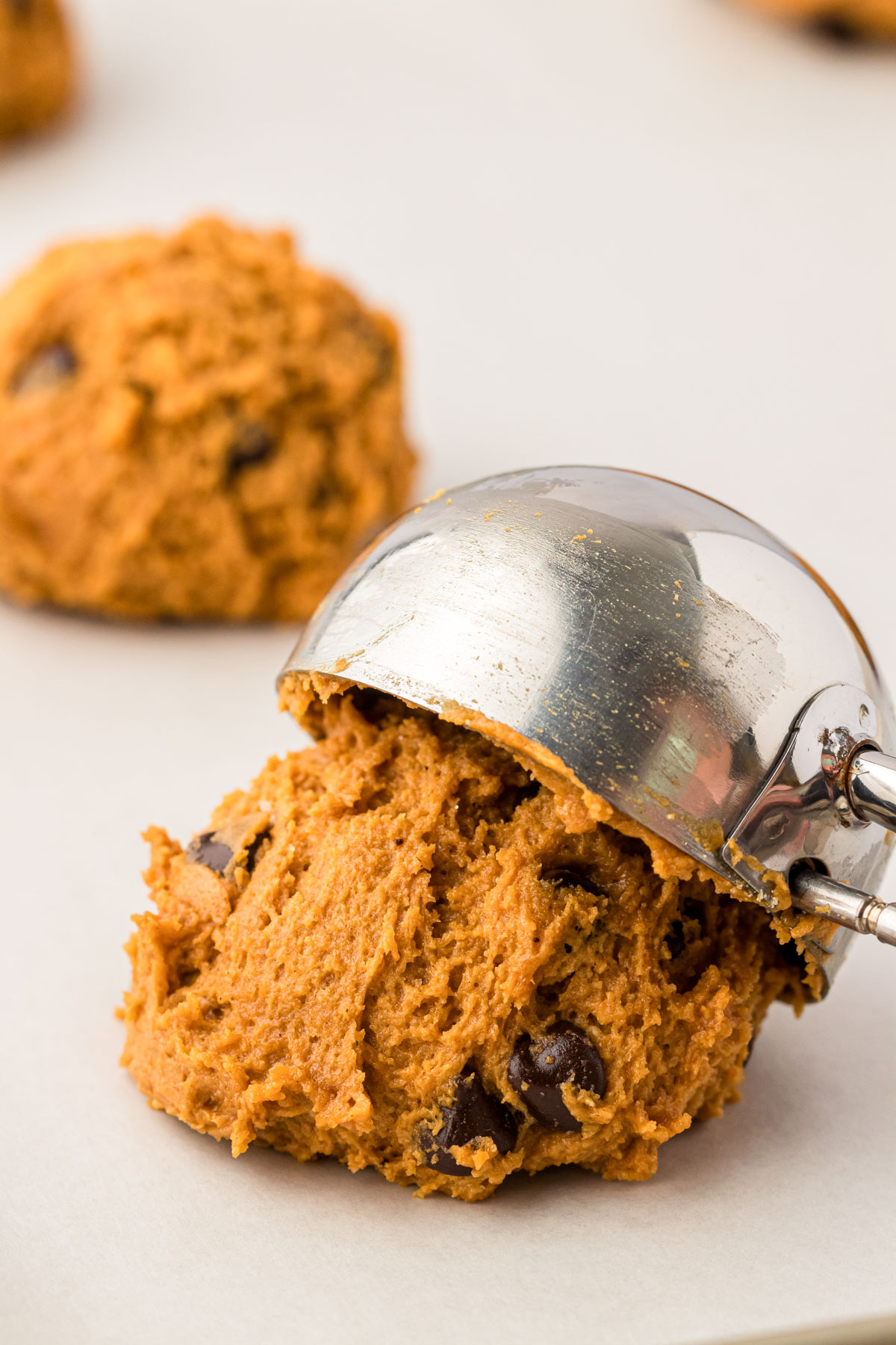 A cookie scoop dropping scoops of pumpkin chocolate chip cookie dough on a parchment lined baking sheet.