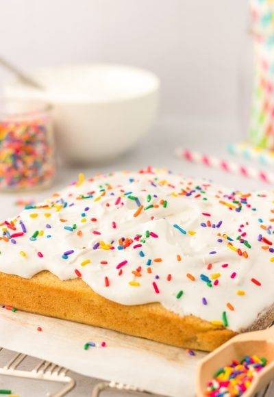 A square vanilla cake on a wire rack that's been frosted and topped with rainbow sprinkles.