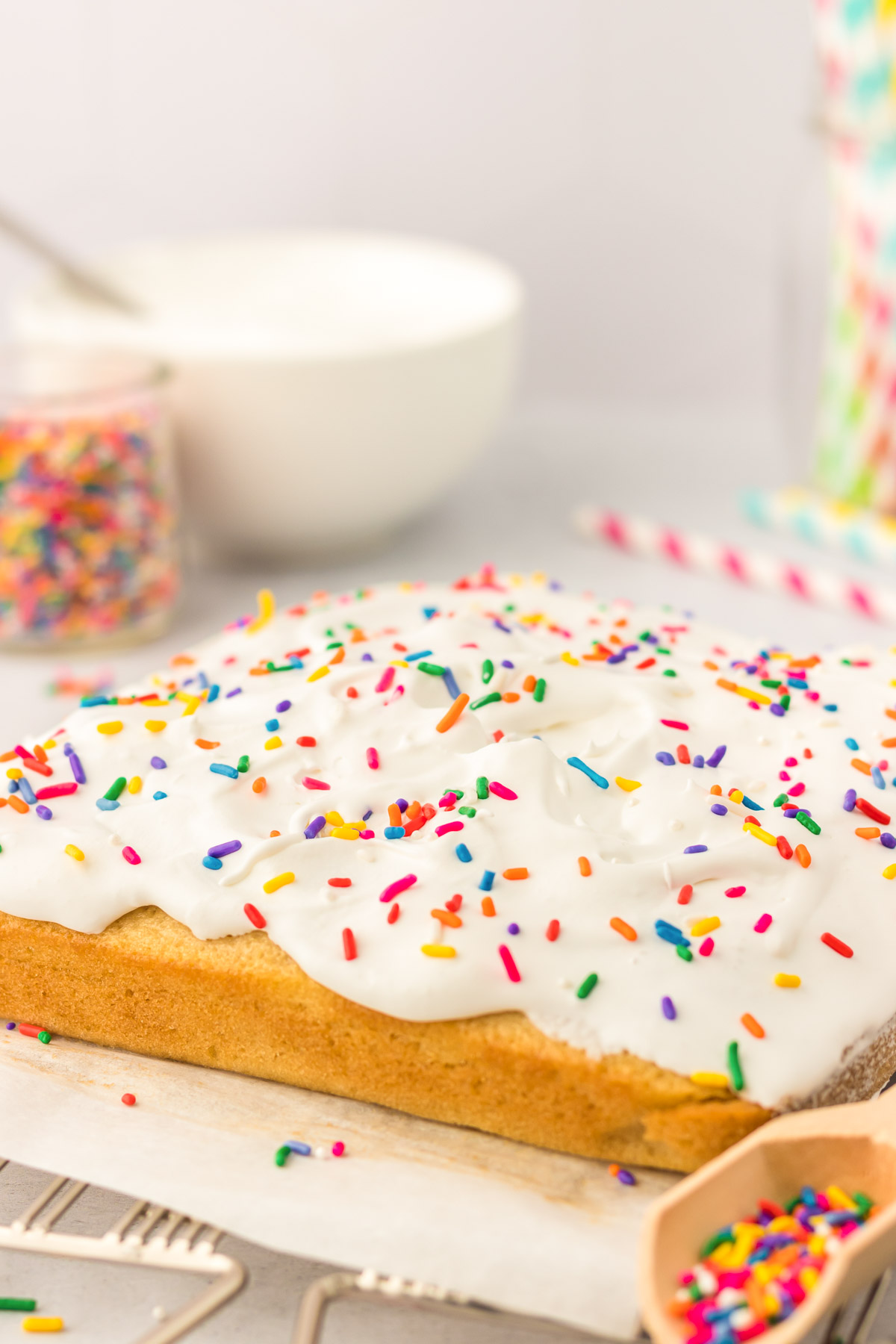 A square vanilla cake on a wire rack that's been frosted and topped with rainbow sprinkles.