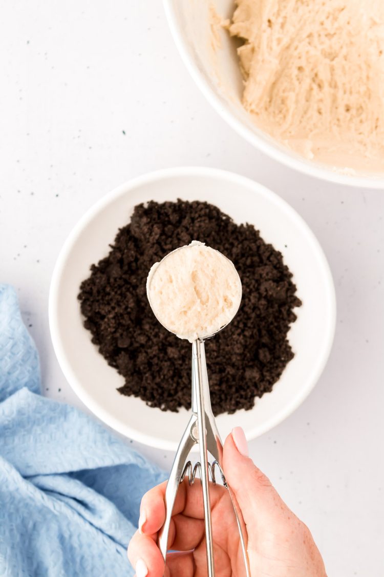 A cookie scoop with sugar cookie dough over a bowl of crushed oreos.