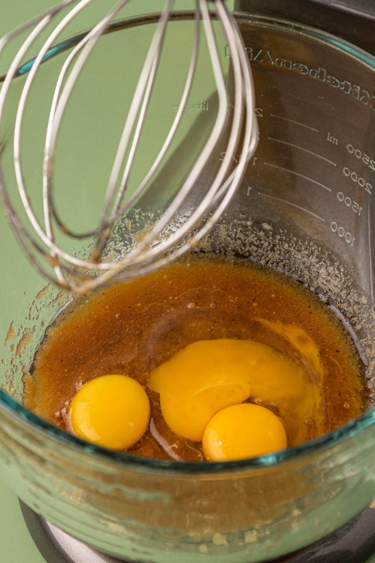 Eggs being added to a mixing bowl with brown butter and sugar.