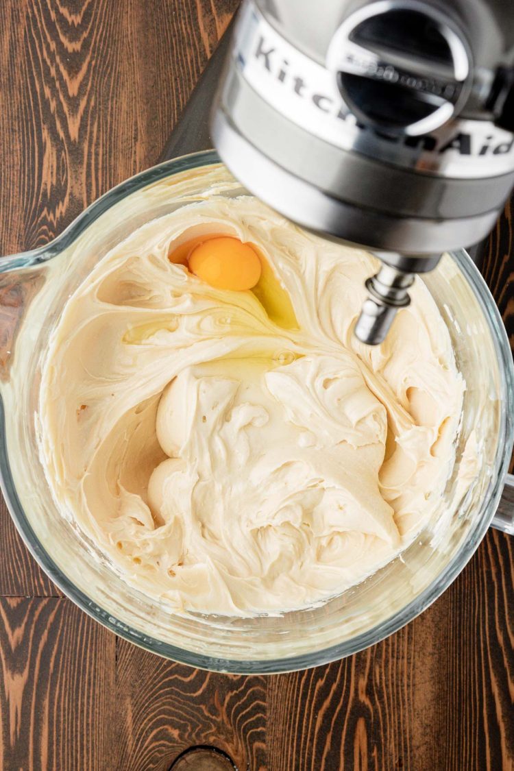 Egg being added to cheesecake filling mix in a stand mixer bowl.