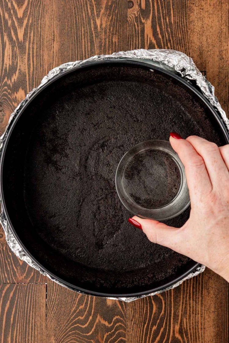 A woman's hand pressing a glass into the bottom of a springform pan filled with oreo crust.