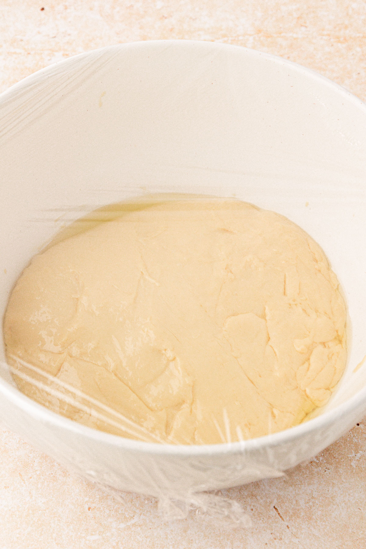 Cinnamon roll dough in a white bowl covered in cling wrap to proof.