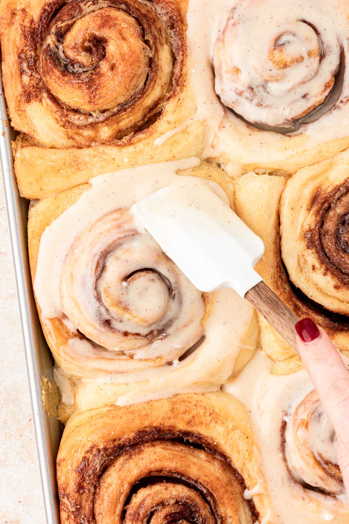 A rubber spatula spreading icing over cinnamon rolls.