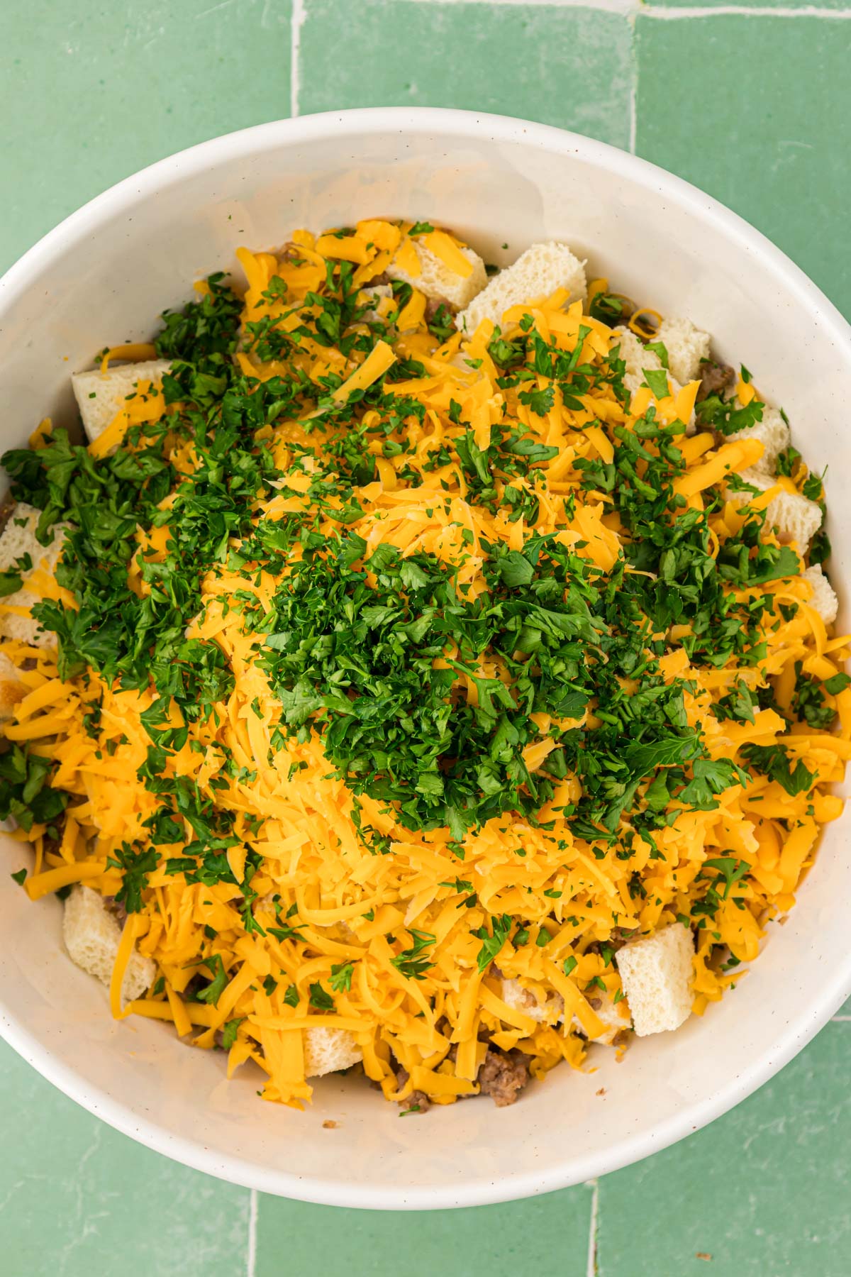 Parsley, cheese, bread, sausage, onions, and spices being mixed together to make a strata mixture.