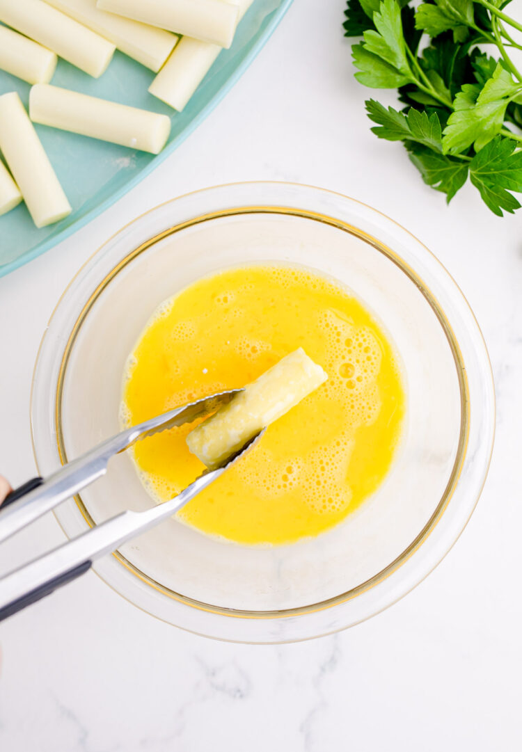 Cheese sticks being dipped in egg wash with tongs.