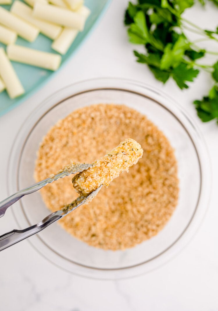 A bread crumb covered mozzarella stick being held over a bowl with tongs.