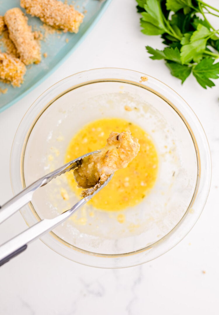 Tongs dipping mozzarella sticks in an eggwash.