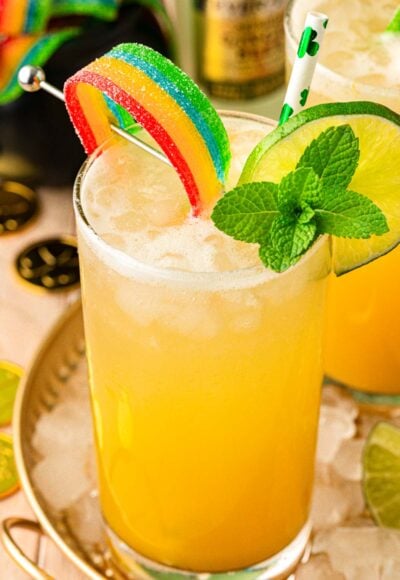 A glass with a yellow drink in it garnished with mint, lime wheel, and rainbow candy.