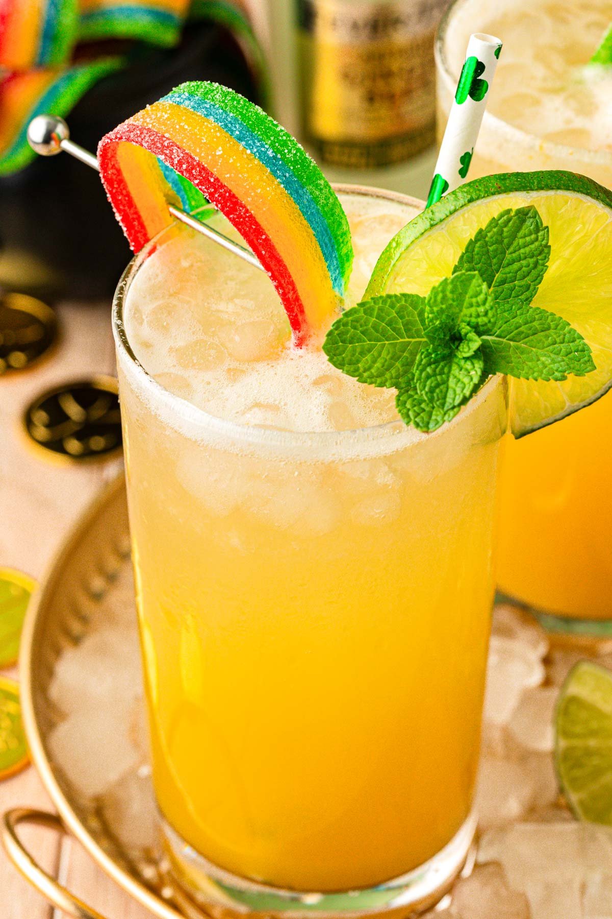 A glass with a yellow drink in it garnished with mint, lime wheel, and rainbow candy.