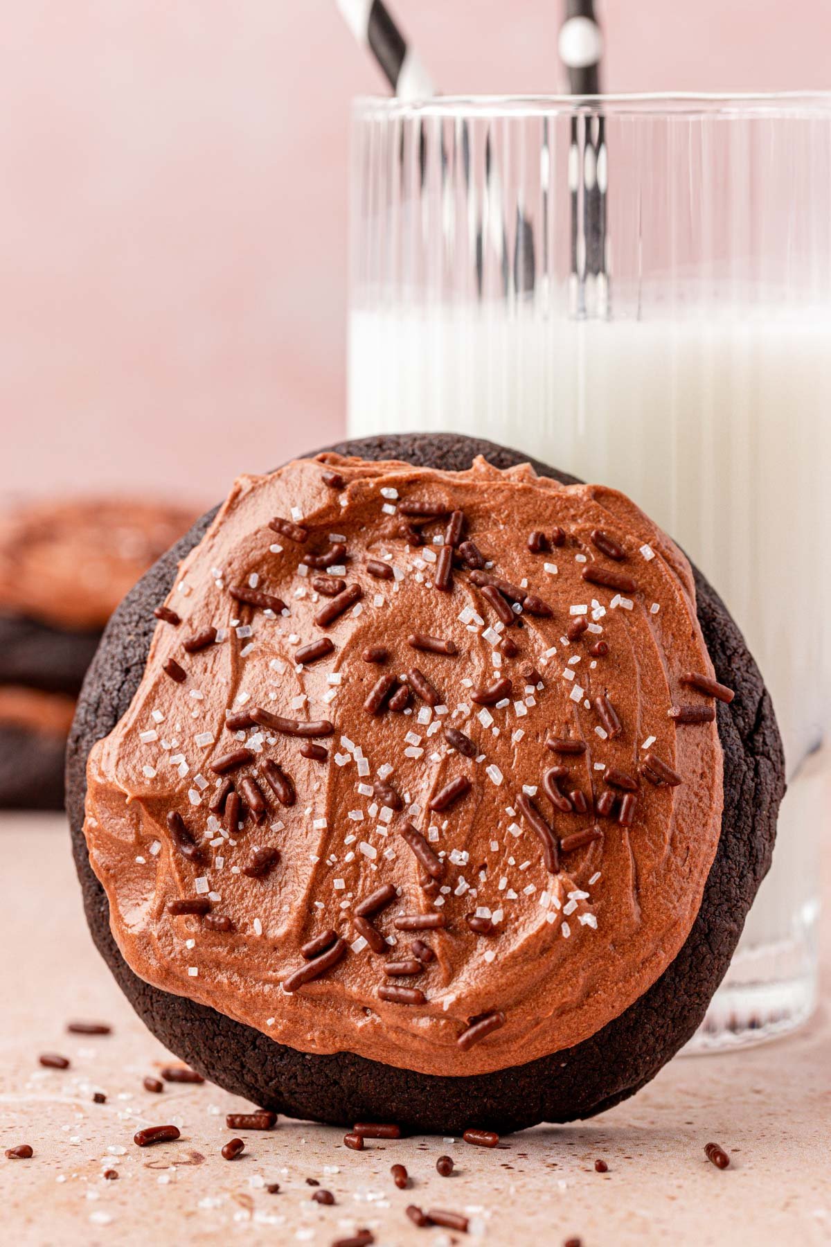 Close up of a big chocolate frosted cookie leaning against a glass of milk.