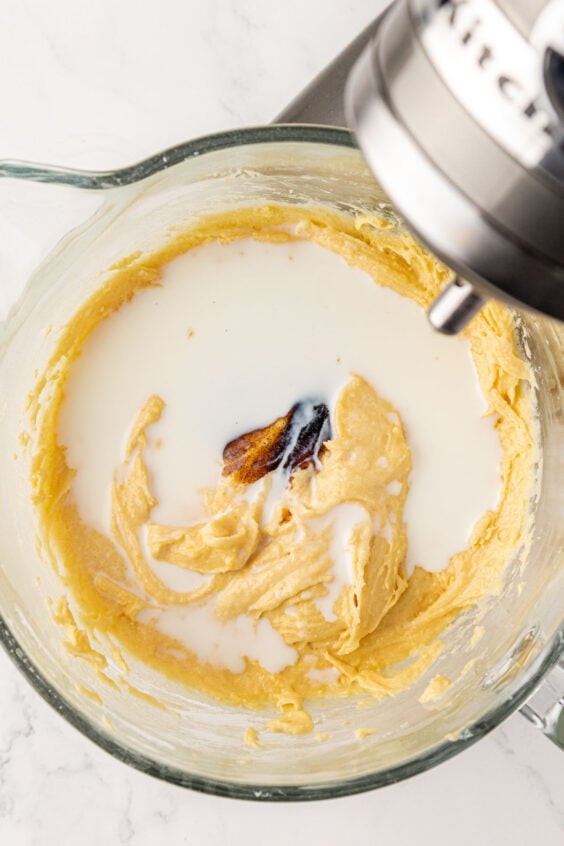 Vanilla bean paste and milk being added to cake batter in a mixing bowl.