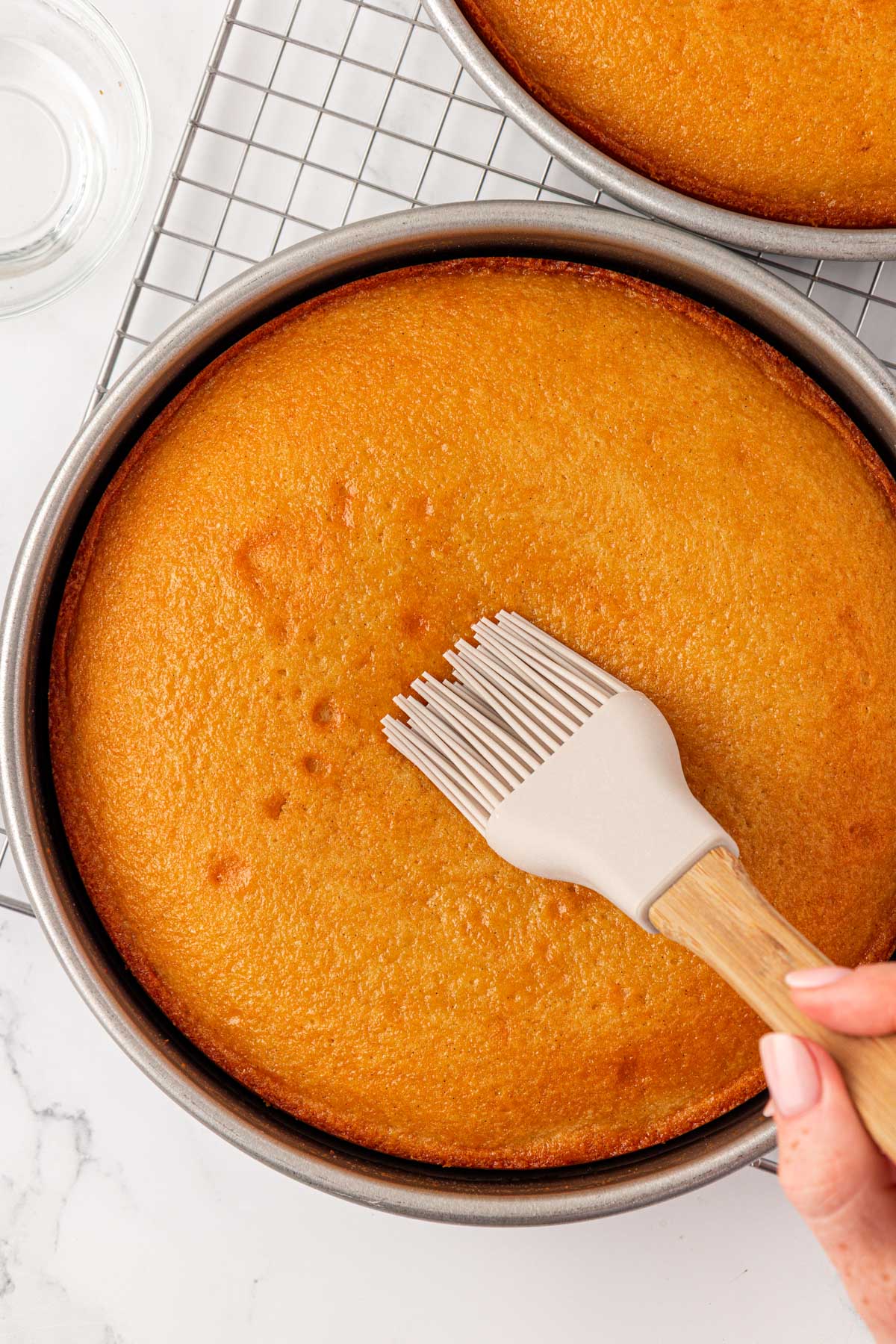 Golden round cake being brush with simple syrup in a pan.