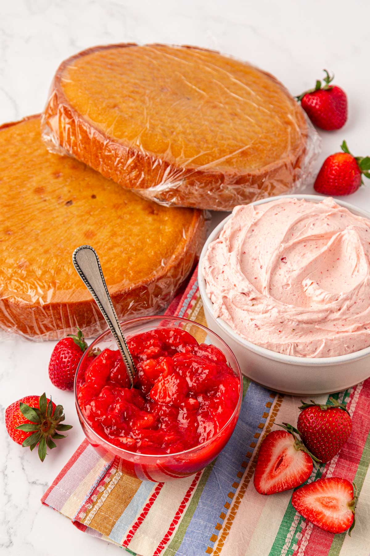 Bowls of strawberry cake filling and strawberry frosting and two round vanilla cakes on a table.