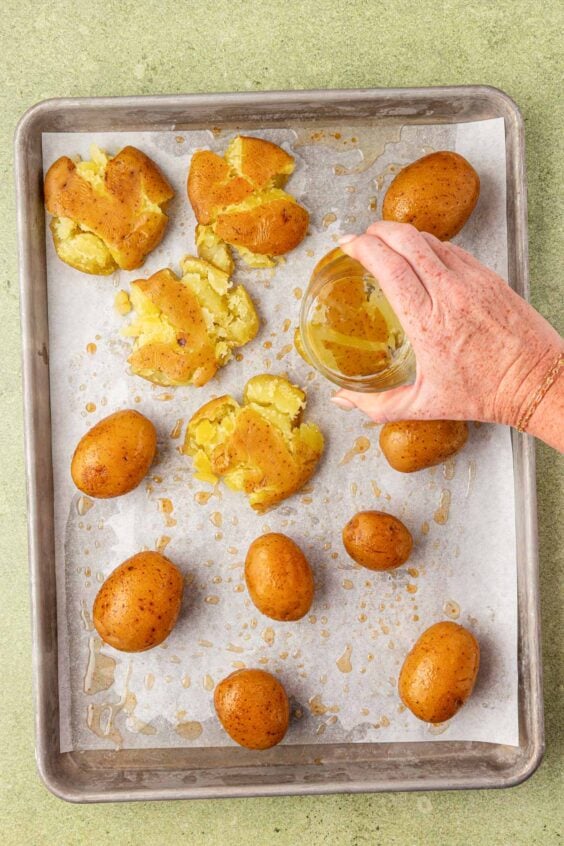 A woman's hand smashing potatoes on a baking pan with the bottom of a glass.