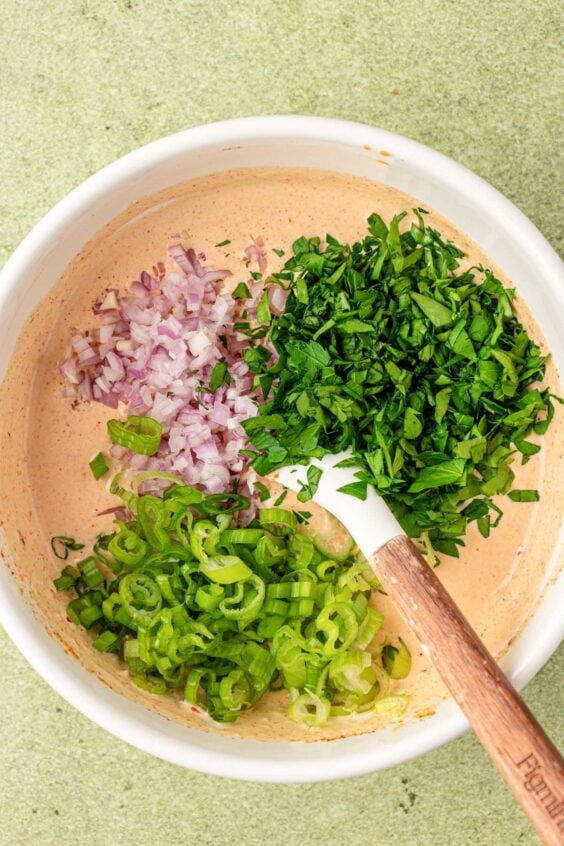 Shallots and herbs being added to a white bowl with a creamy dressing.