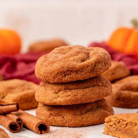 A stack of three pumpkin snickerdoodle cookies on a table with more cookies and cinnamon sticks around them.